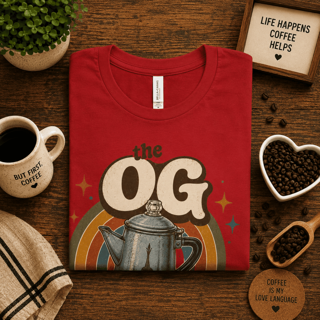 Folded mock up of The OG graphic t-shirt in red, surrounded coffee beans, a coffee scoop and a small house plant.