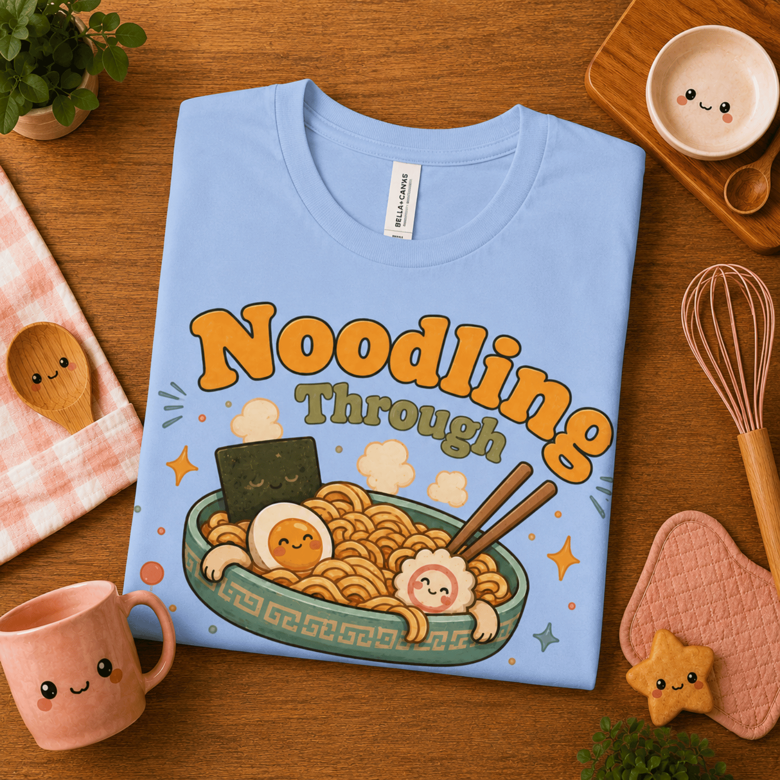 Folded mock up of Noodling Through Life graphic t-shirt in baby blue, surrounded by small kitchen utensils.