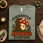 A folded mockup of Currently Avoiding funny mushroom graphic t-shirt in sage.