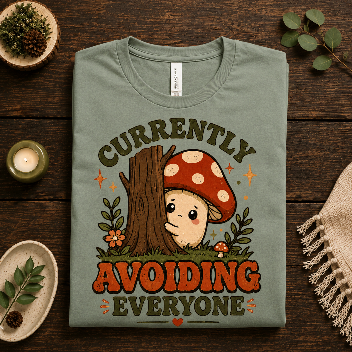 A folded mockup of Currently Avoiding funny mushroom graphic t-shirt in sage.