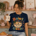 Woman wearing Bowl Goals graphic t-shirt in navy sitting on kitchen stool, drinking coffee.