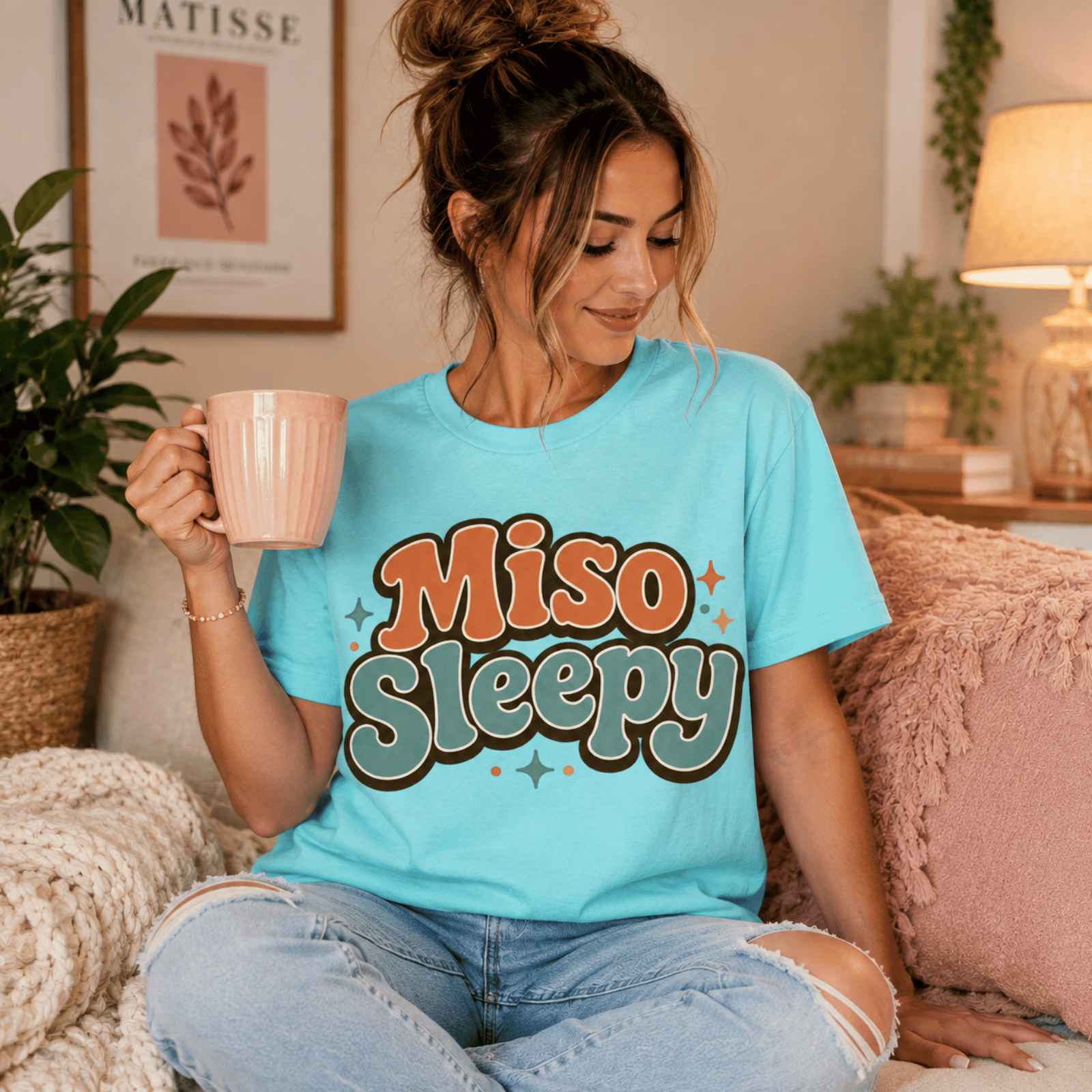 Woman wearing Miso Sleepy graphic t-shirt in aqua sitting on couch drinking coffee.