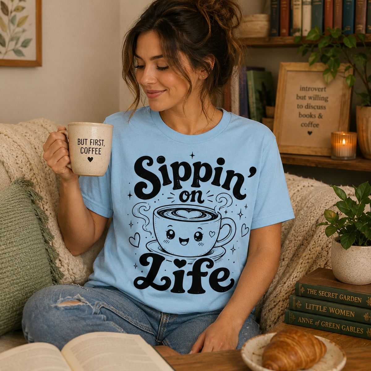 Woman drinking coffee in her living room wearing a baby blue Sippin' On Life coffee graphic t-shirt.