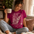 A woman wearing a berry Cat Observing graphic t-shirt drinking coffee.