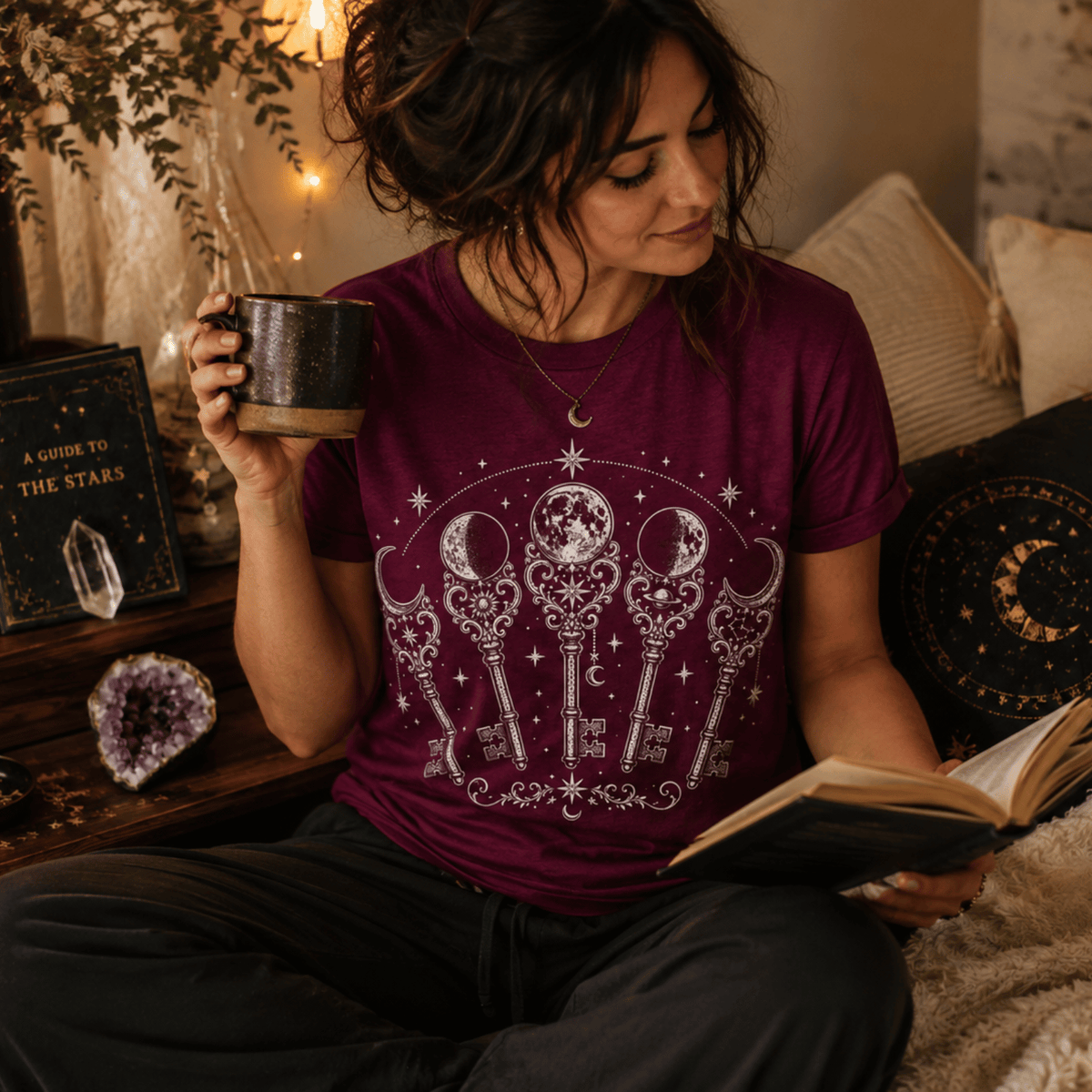 A woman wearing a maroon Moon Phase Keys celestial graphic t-shirt reading a book.