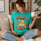 A woman wearing a heather sea green Floating Through Life graphic t-shirt drinking coffee.