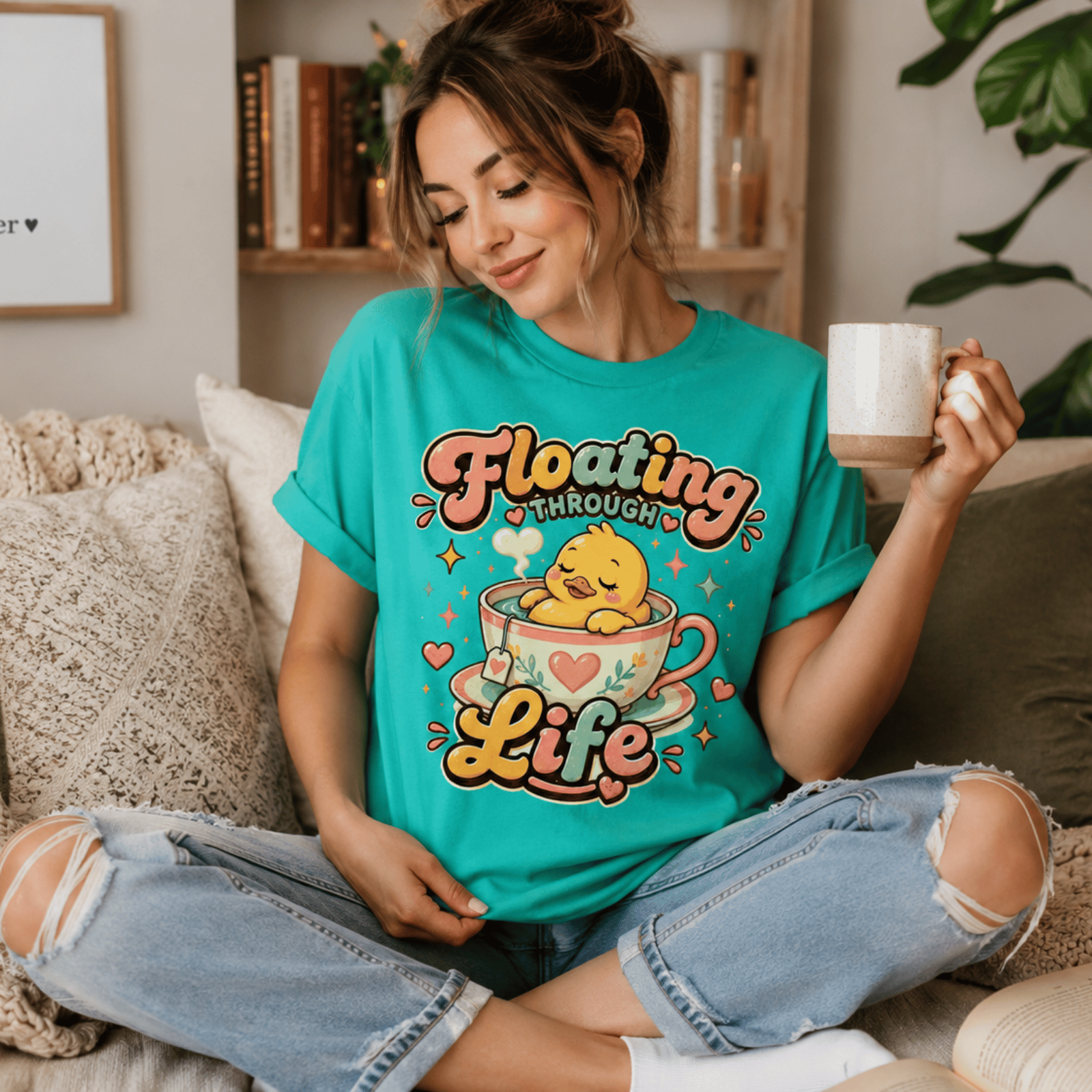 A woman wearing a heather sea green Floating Through Life graphic t-shirt drinking coffee.