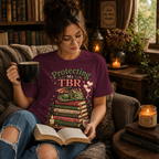 A woman wearing a maroon Protecting My TBR cute book graphic t-shirt drinking coffee and reading a book.