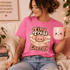 Woman wearing Tiny Treat Energy graphic t-shirt in charity pink sitting on couch drinking coffee.