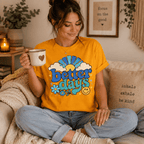 Woman sitting cross-legged on a couch wearing the Better Days Premium T-Shirt in gold, holding a mug