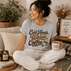 Woman sitting on a couch holding a mug, wearing the Cold Hands, Warm Coffee Premium T-Shirt in athletic heather gray