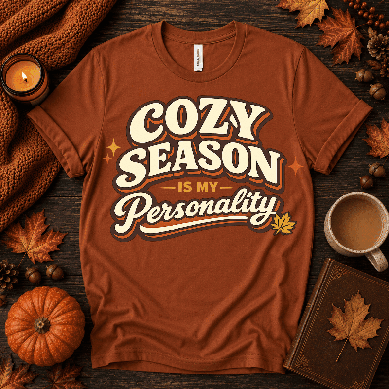 Cozy Season Premium T-Shirt in autumn rust orange, flat lay with "Cozy Season Is My Personality" script and maple leaf graphic, styled with fall props