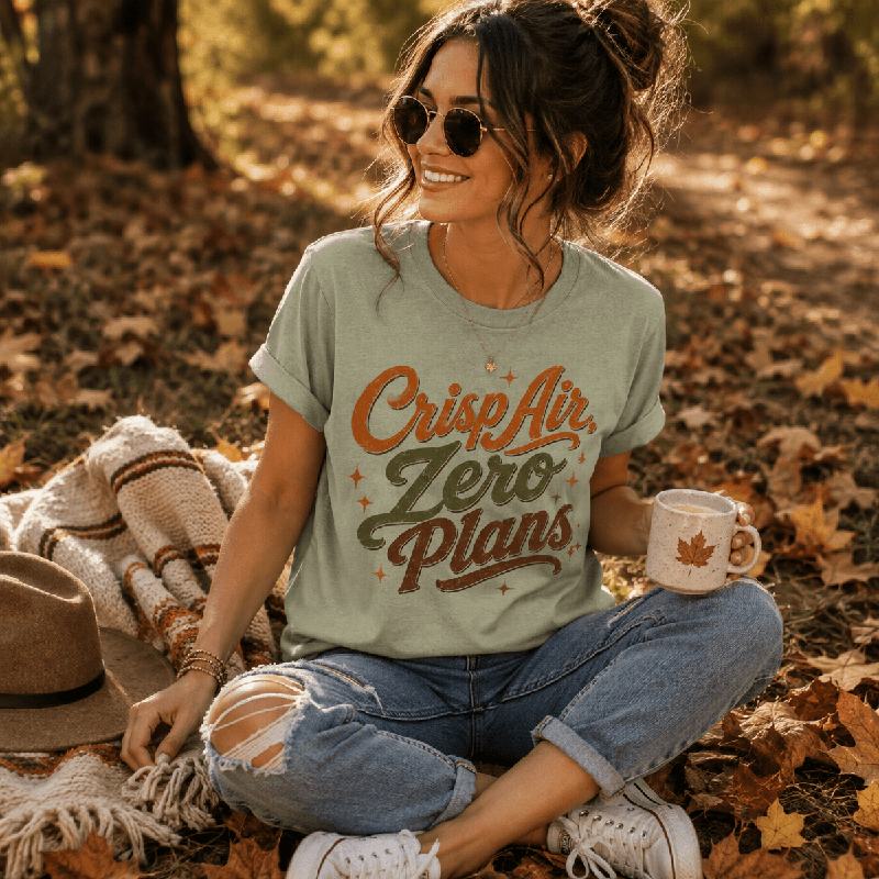 Woman sitting outdoors on fallen leaves holding a mug, wearing the Crisp Air Premium T-Shirt in sage