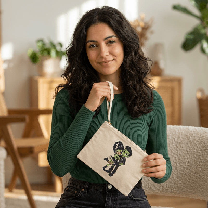 Woman holding the natural canvas Frankie & Frannie Accessory Pouch by its wrist strap, printed with a chibi Frankenstein couple graphic
